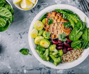 Healthy buddha bowl lunch with grilled chicken, quinoa, spinach, avocado, brussels sprouts, broccoli, red beans with sesame seeds on dark gray background. Top view.
